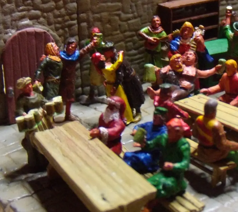 A photo of a miniature diorama showing a medieval tavern scene with painted figurines. Several figures are seated at wooden…