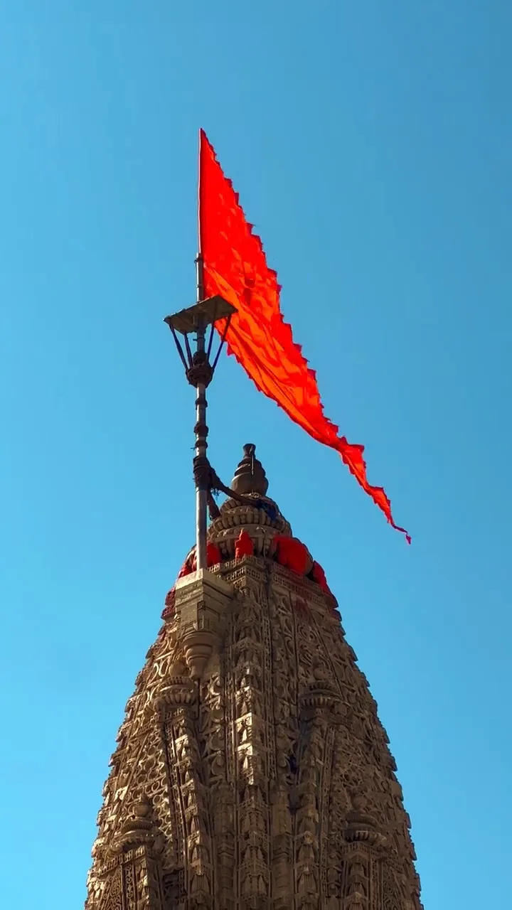 A low-angle shot shows the ornate, intricately carved spire of a temple against a clear blue sky, topped by a bright red…
