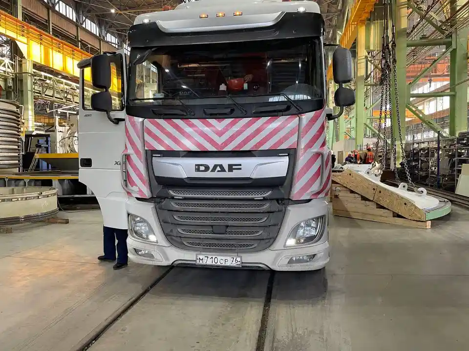 A white DAF truck with red and white diagonal hazard stripes on its front grille is parked inside a large industrial…