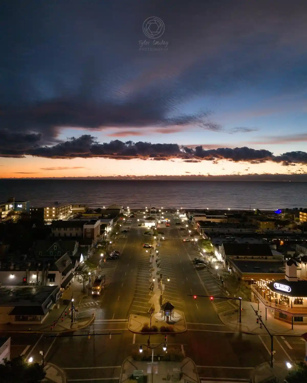 An aerial photo of a coastal town at dusk, showing a wide street with parked cars, illuminated buildings, and a dramatic sky…