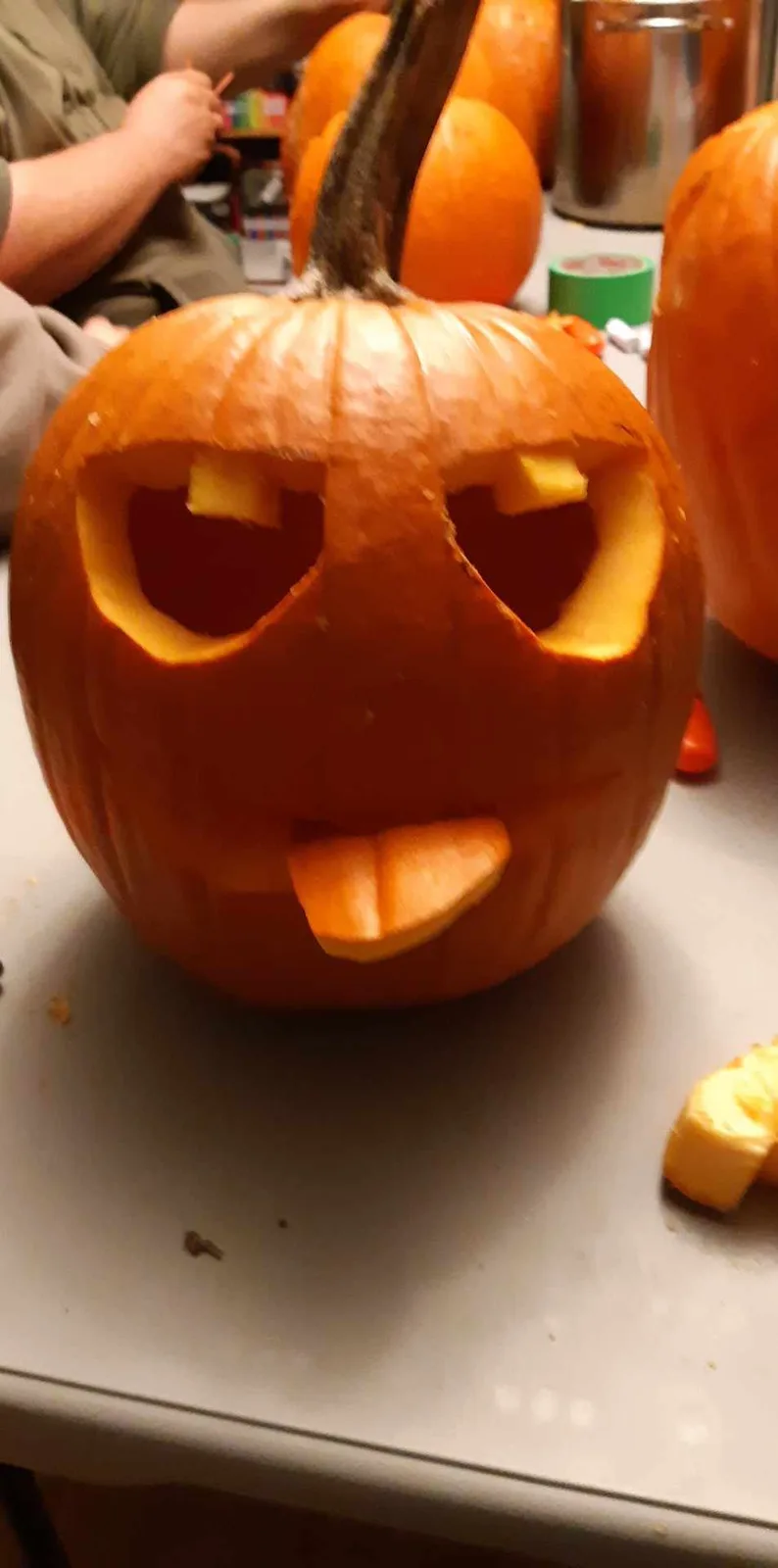 A close-up photo of a carved pumpkin with a goofy face, featuring large eye sockets with small yellow pieces of pumpkin…
