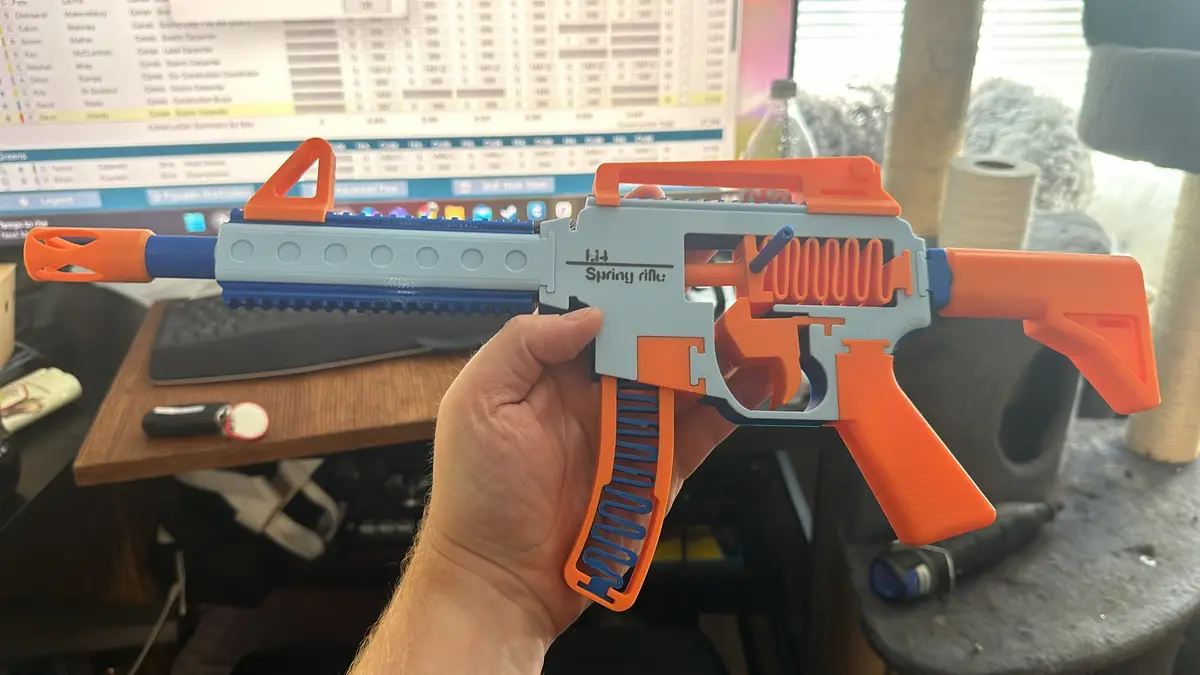 A hand holds a disassembled toy gun with orange and blue plastic parts, revealing its internal spring mechanism. The text…