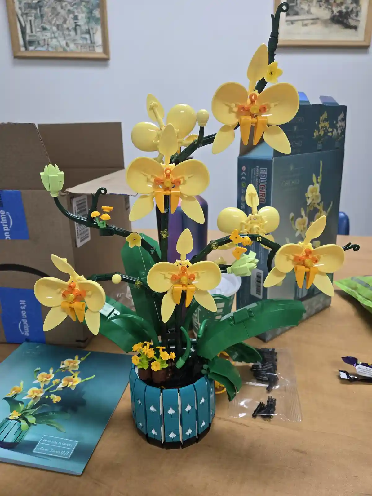 A completed LEGO model of a yellow orchid plant with green stems and leaves sits on a wooden table, surrounded by its…