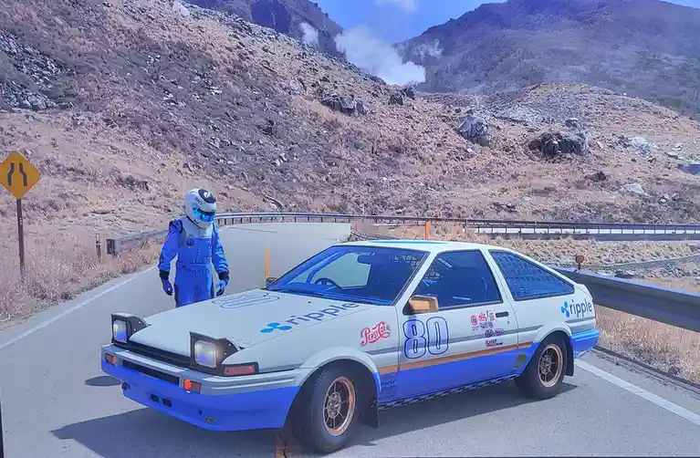 A person in a blue racing suit and helmet stands beside a white and blue Toyota AE86 race car with 'ripple' and 'pepsi cola'…