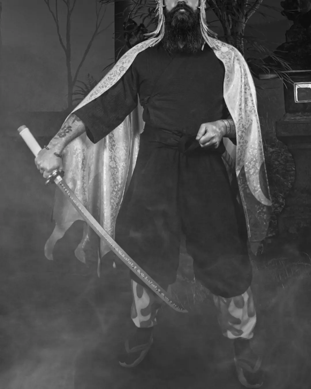 A black-and-white photo shows a man with a full beard and long hair, dressed in traditional samurai attire including a…