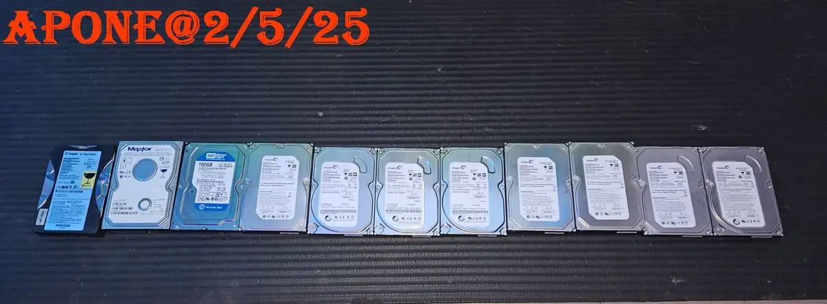 A row of ten computer hard drives of various brands and sizes lies on a dark textured surface. The text 'APONE@2/5/25' is…