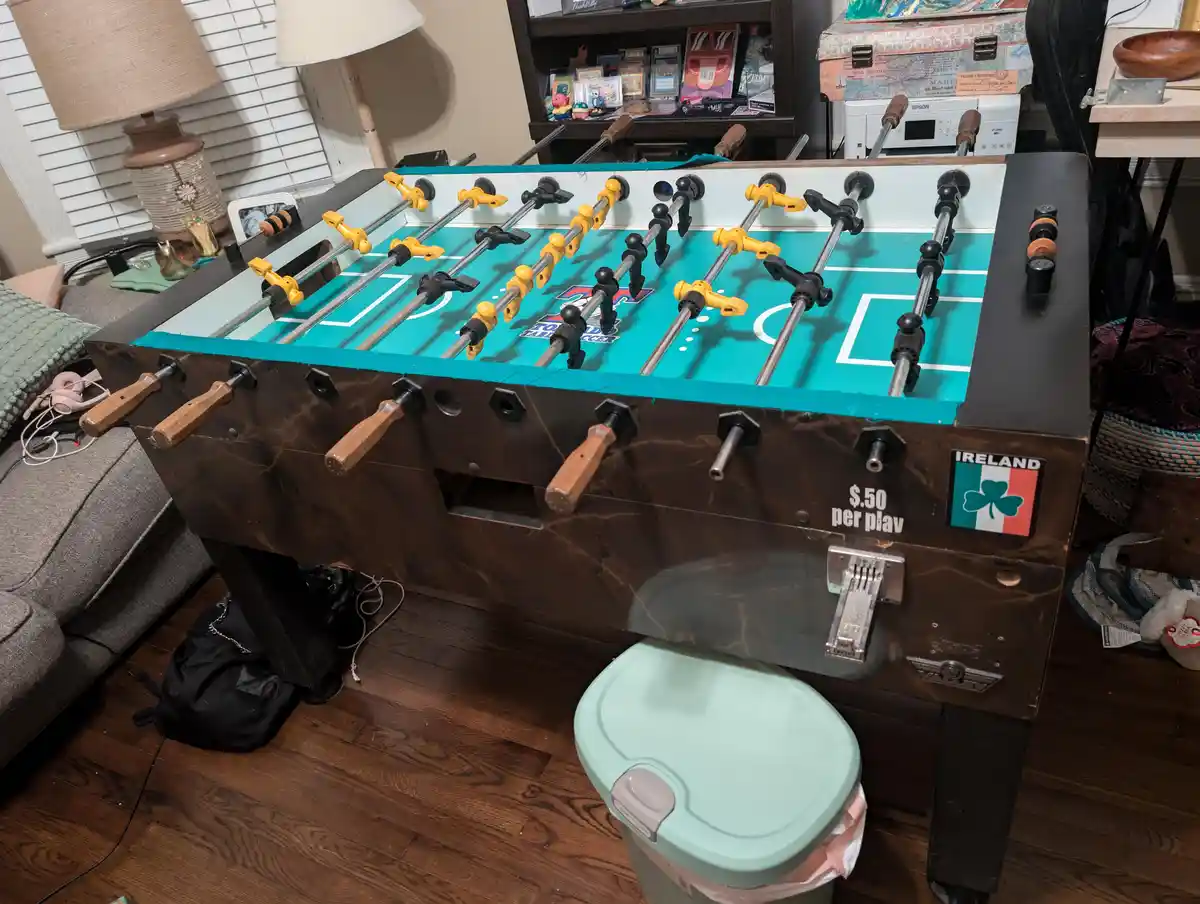 A foosball table with a brown marble-patterned frame and a teal playing surface is shown in a living room. It has wooden…