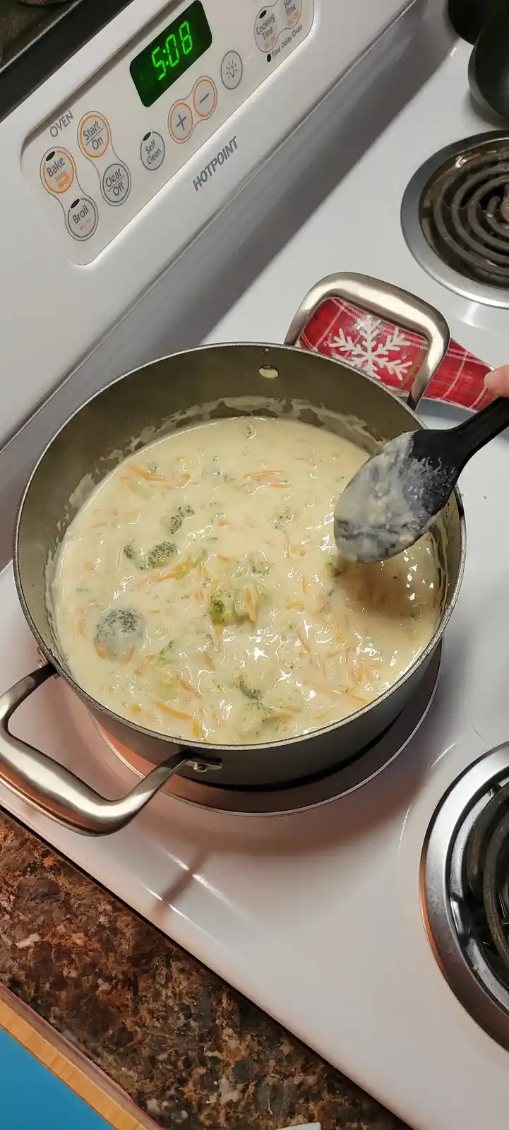 A close-up photo shows a pot of creamy broccoli and cheese soup simmering on a white Hotpoint stovetop. A black spoon lifts…