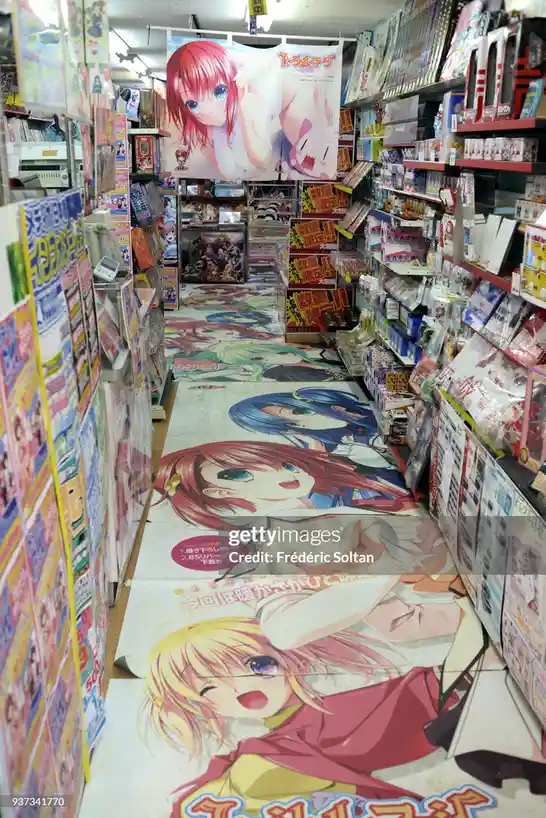 This image shows a narrow aisle in a Japanese anime store, where the floor is entirely covered by large, colorful anime…