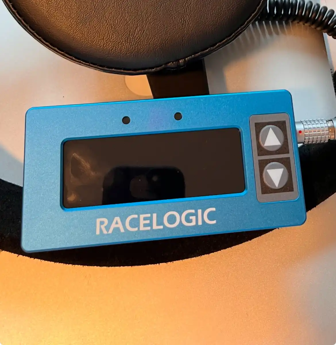 A blue RaceLogic device with a blank screen and up/down arrow buttons sits on a black padded surface, with a coiled cable…