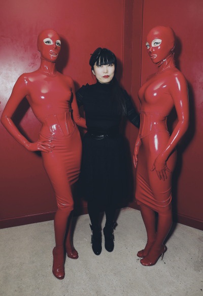 Three people stand in a corner against a red wall; two are in full-body shiny red latex suits with masks, and one in the…