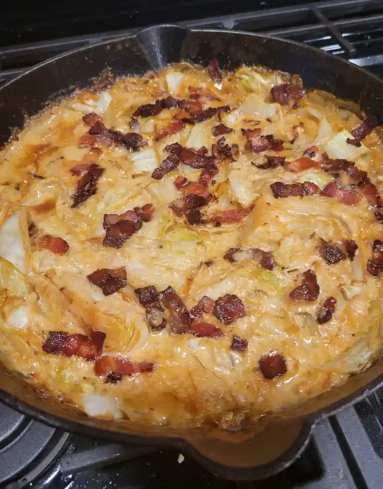 A close-up photo shows a cast-iron skillet on a stovetop containing a cheesy, baked dish with visible cabbage and crispy…