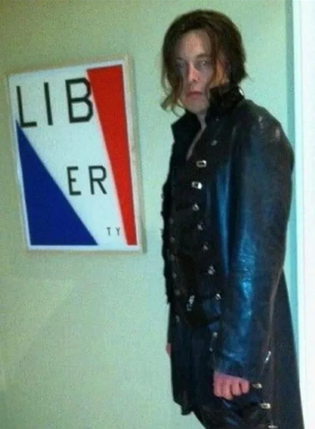 A person with long hair wearing a dark, shiny, buttoned coat stands beside a framed sign that reads 'LIB ER TY' with red and…