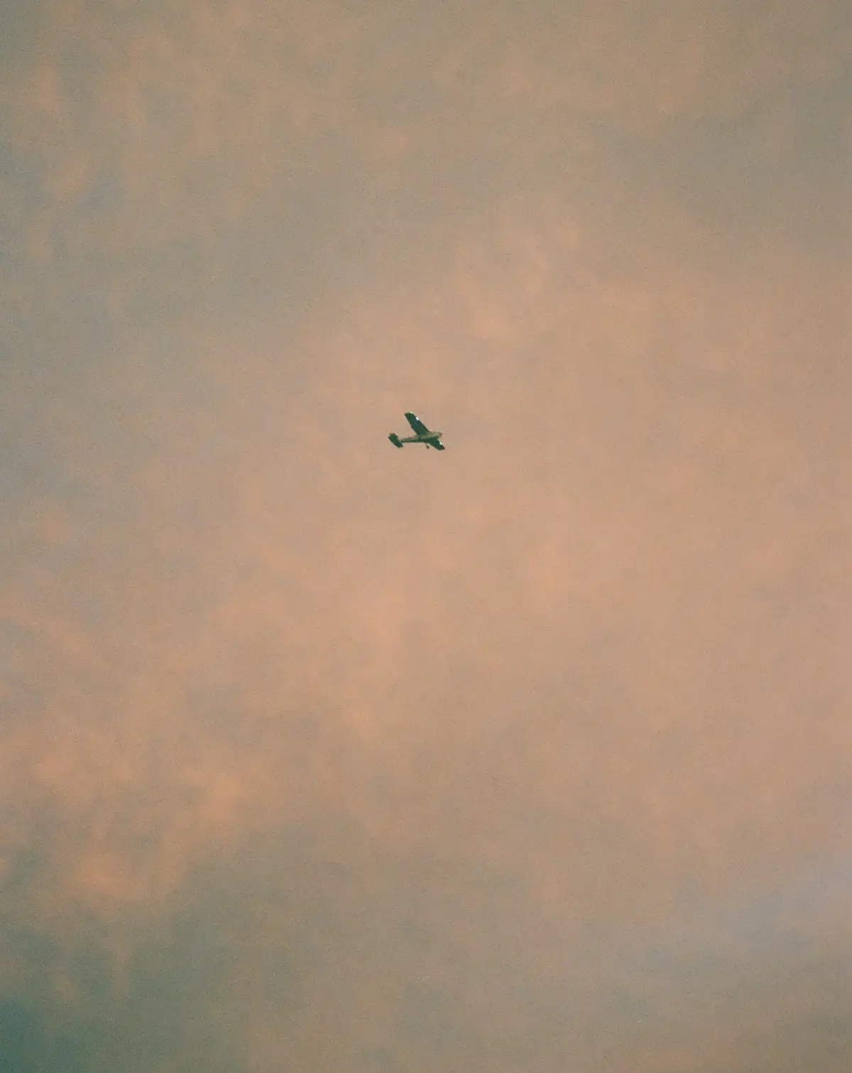 A small, dark-colored airplane is flying alone against a vast, soft pink and orange sky filled with wispy clouds. The plane…