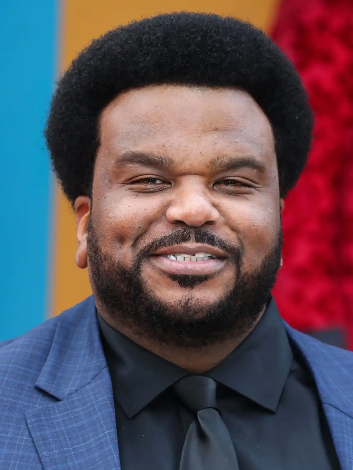 A close-up photo of a smiling man with a full beard and afro hairstyle, wearing a blue plaid suit jacket over a black shirt…