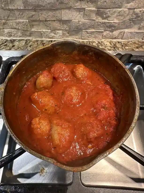 A close-up photo shows a large, well-used metal pot on a stovetop, filled with red tomato sauce and several round meatballs…