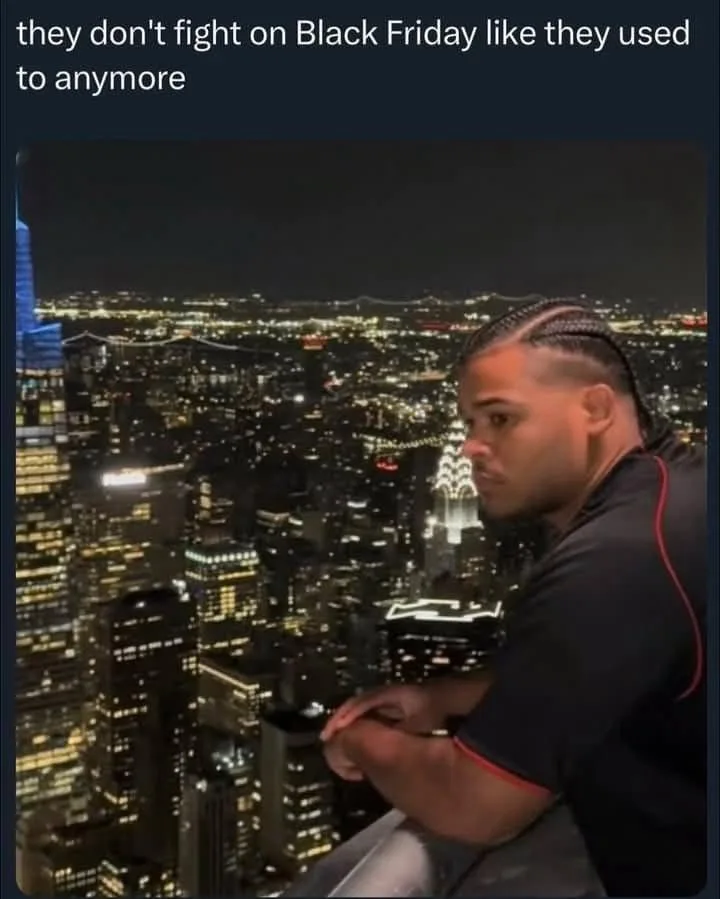 A man with braided hair sits on a high vantage point overlooking a brightly lit cityscape at night, looking off to the side…