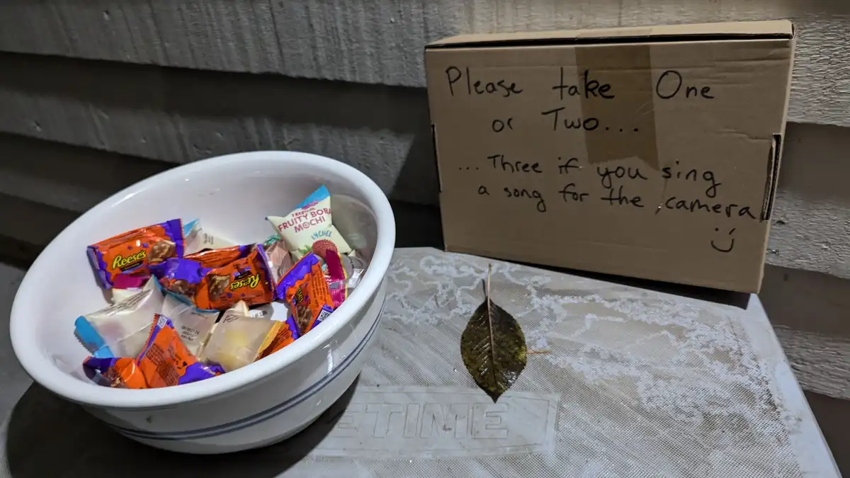 A white ceramic bowl filled with assorted wrapped candies sits next to a cardboard box with handwritten text. The text reads…