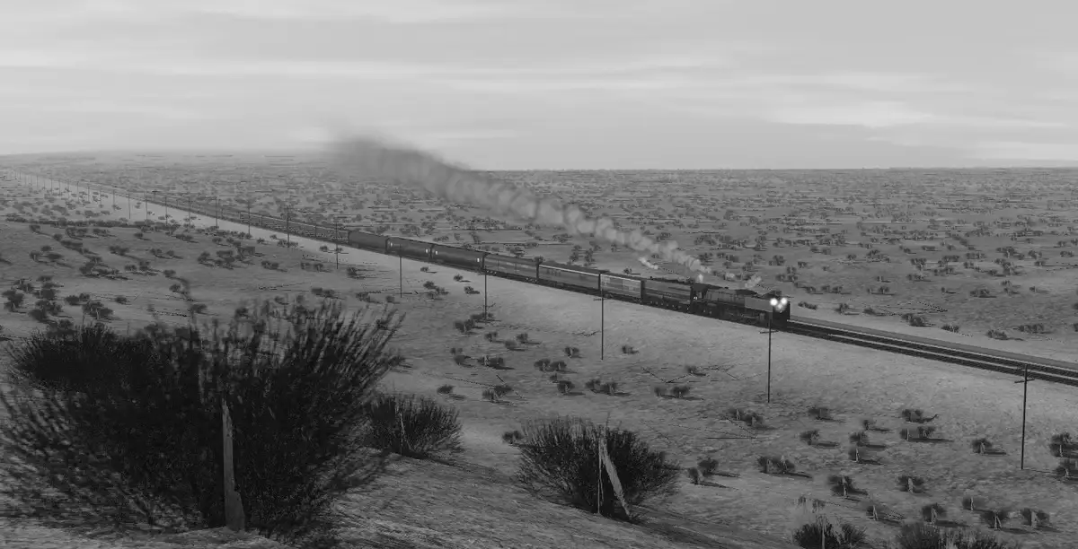 A black-and-white image shows a long steam train traveling through a vast, arid desert landscape, emitting smoke as it moves…