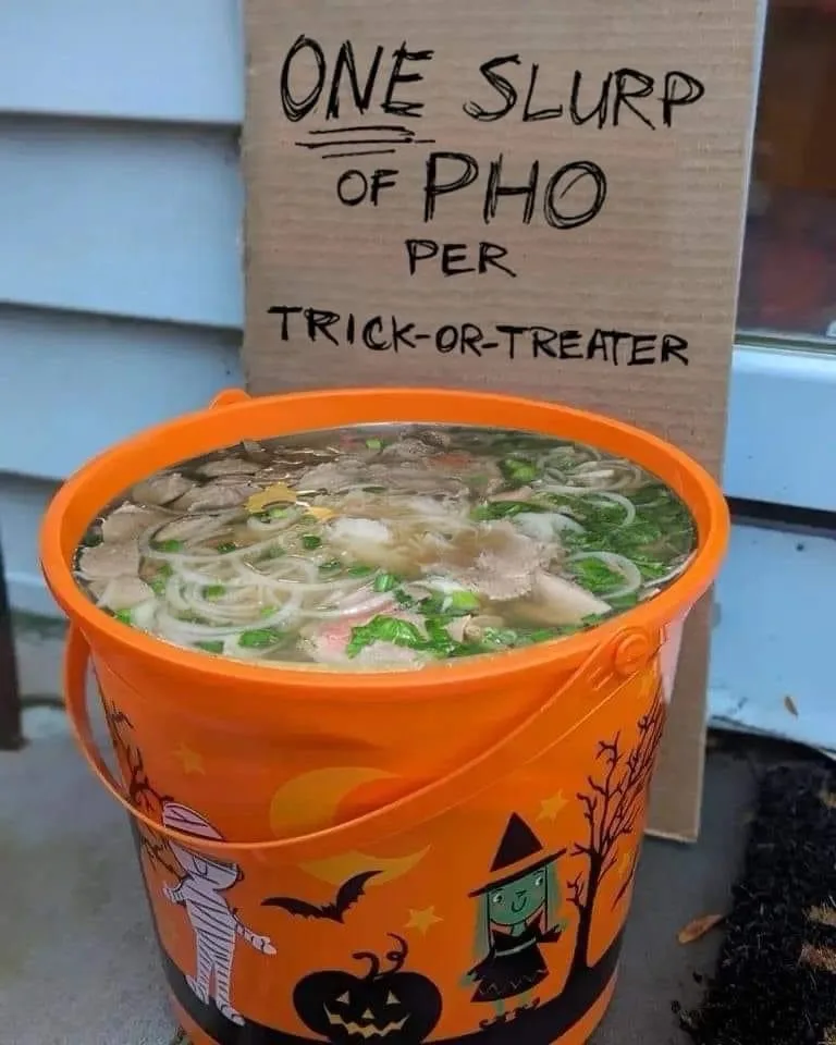 An orange Halloween-themed bucket filled with pho soup sits in front of a cardboard sign that reads 'ONE SLURP OF PHO PER…
