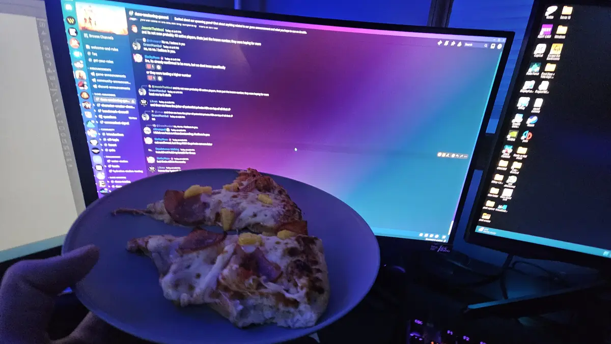 A hand holds a blue plate with two slices of pizza topped with pineapple and bacon in front of a computer monitor displaying…