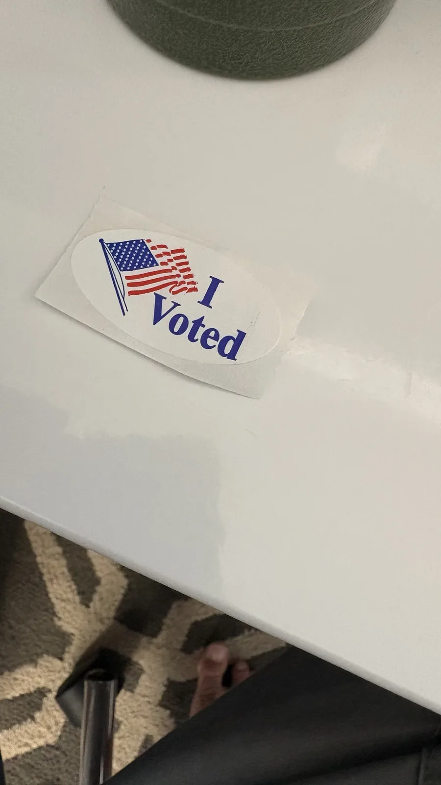A close-up photo shows a slightly crumpled 'I Voted' sticker with an American flag design, stuck on a white surface, likely…