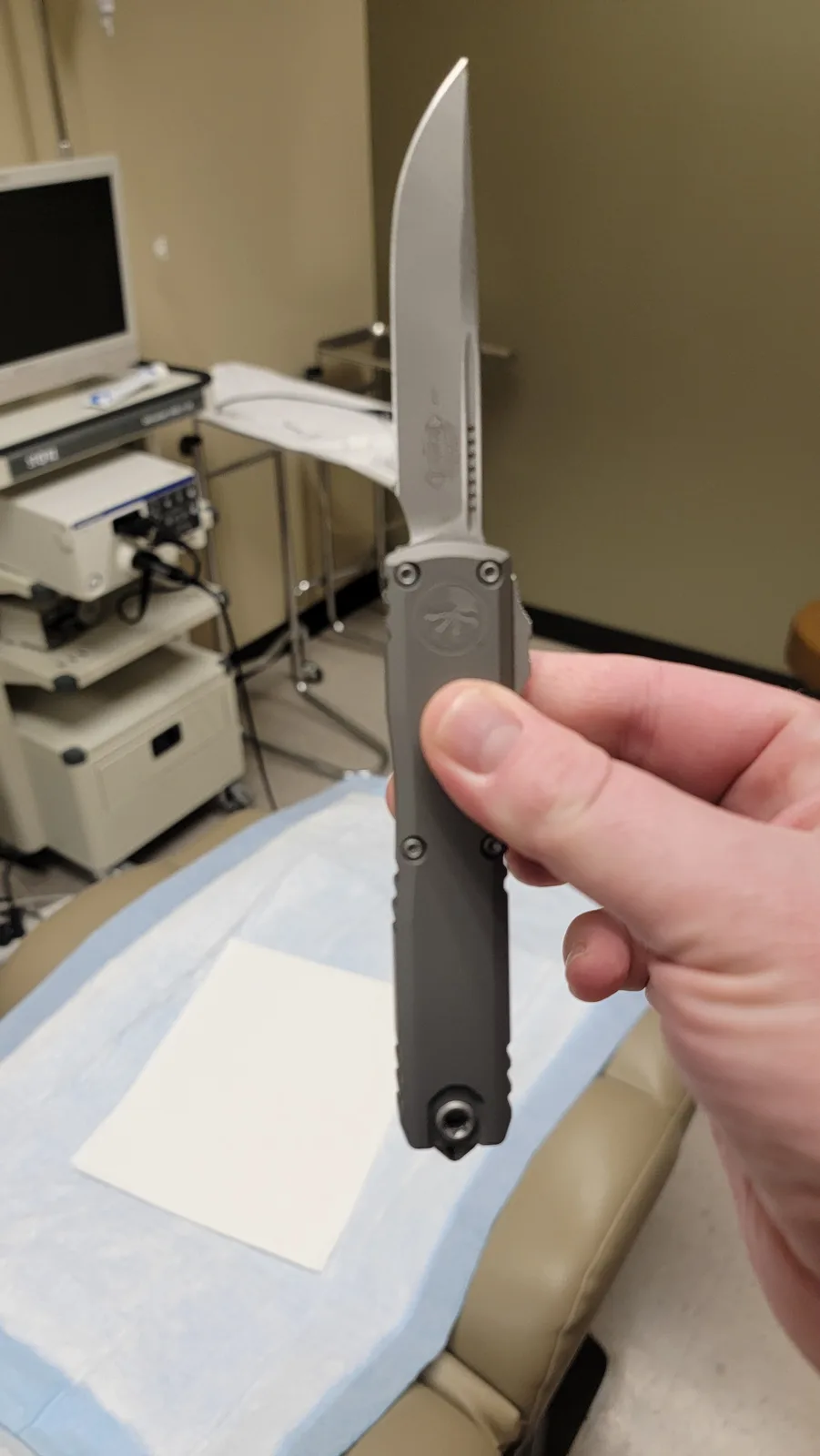 A hand holds a large, gray tactical-style folding knife with a sharp blade, positioned in front of a dental chair covered…
