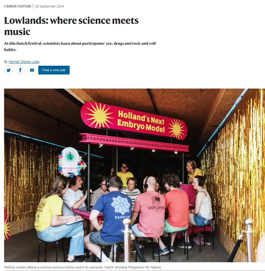 A group of people sit around a table at a science festival booth under a red sign that reads 'Holland's Next Embryo Model'…