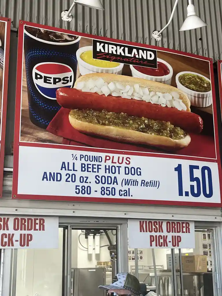 A large advertisement sign for Kirkland Signature shows a hot dog with onions and relish, alongside a Pepsi soda, priced at…