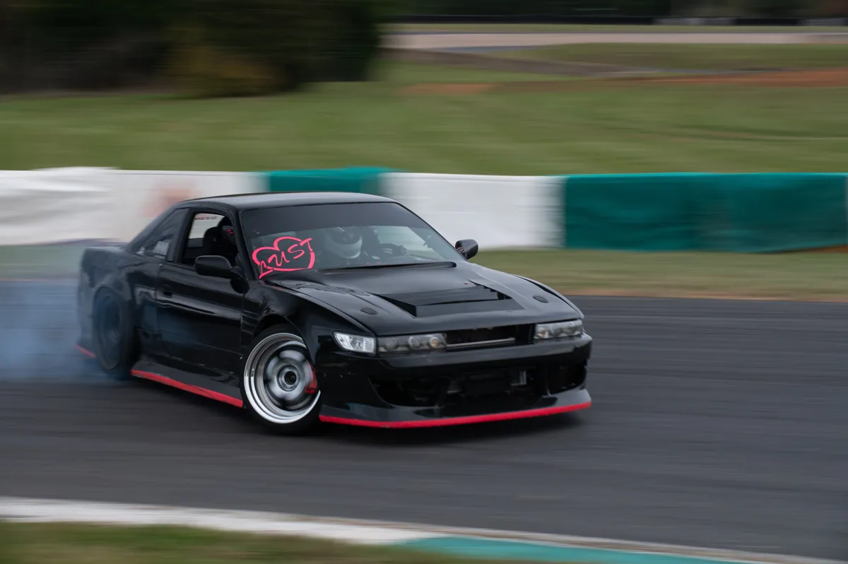 A black Nissan S14 drift car with red trim and a pink 'lust' decal on the windshield is drifting on a racetrack, kicking up…