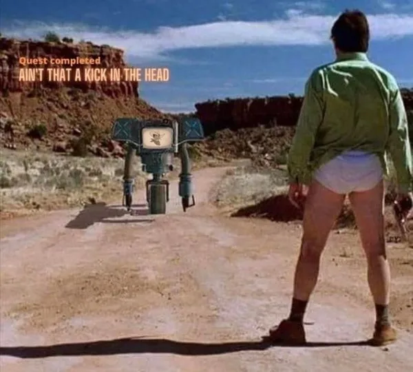 A man in a green shirt and white underwear stands facing a small robot in a desert landscape. The robot has a screen on its…