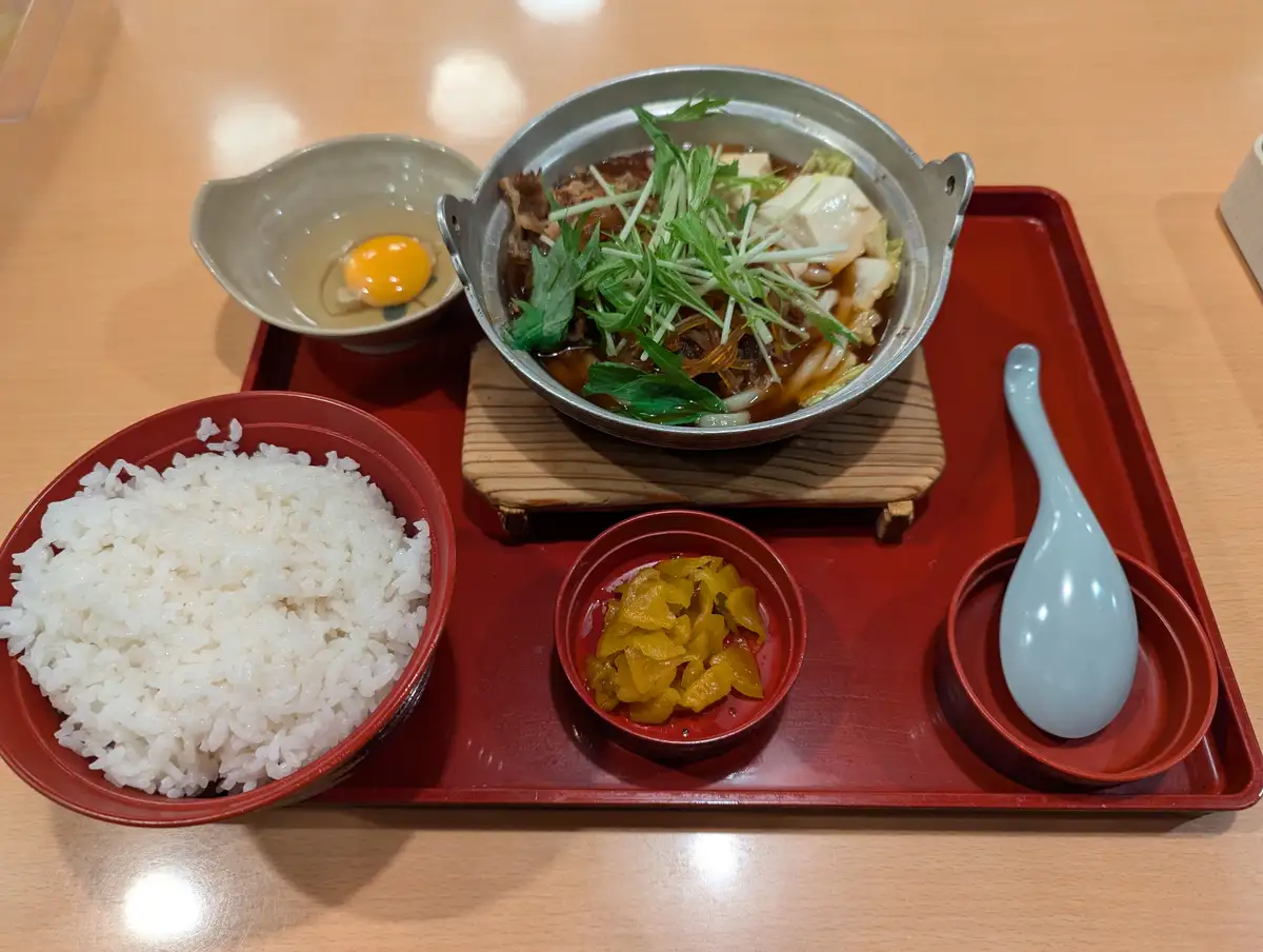 a japanese meal served on a red tray includes a bowl of white rice, a metal pot of hot soup with tofu, greens, and noodles…