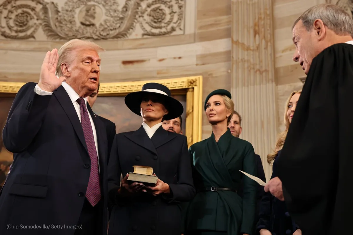 A man in a dark suit raises his right hand while standing next to a woman holding a book, with another woman in a green…