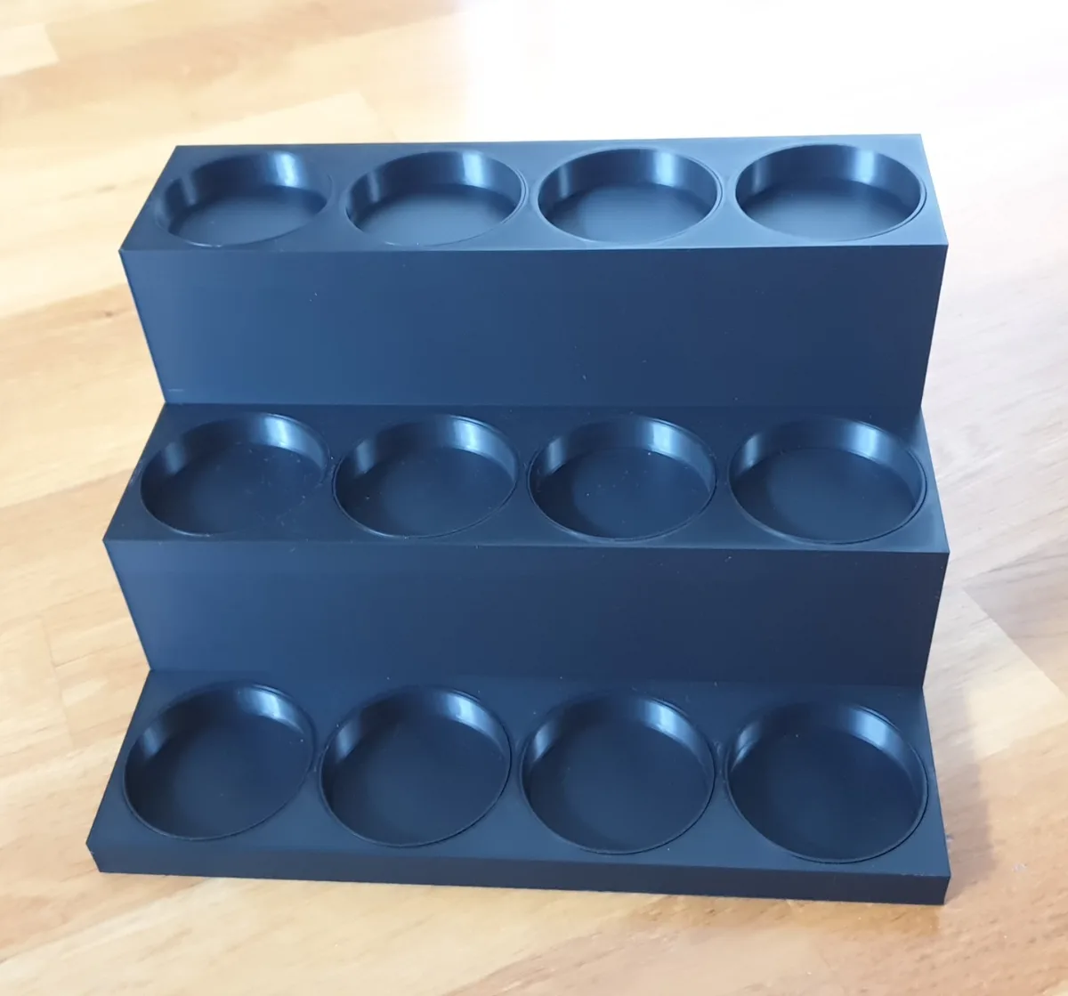 A black, three-tiered, rectangular holder with circular indentations sits on a light wooden floor. Each tier has four…