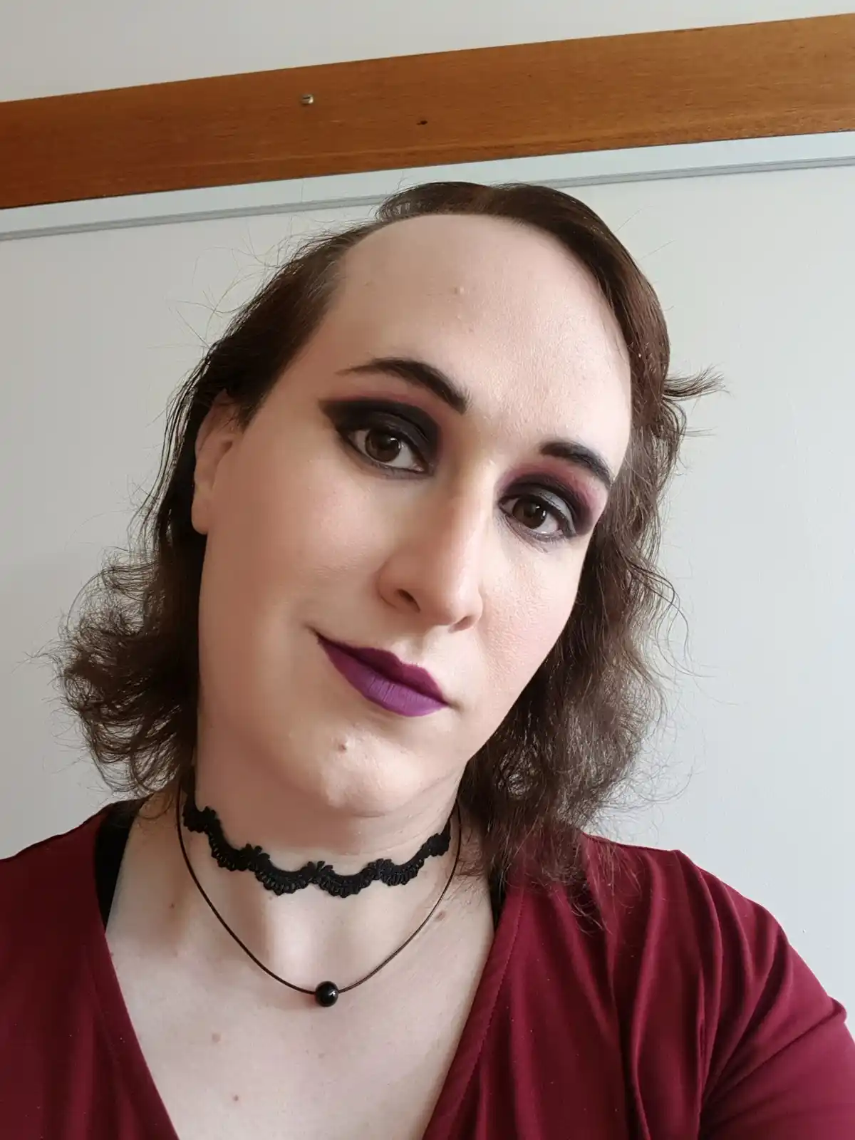 A person with dark, dramatic eye makeup, purple lipstick, and a black lace choker looks directly at the camera with a tired…
