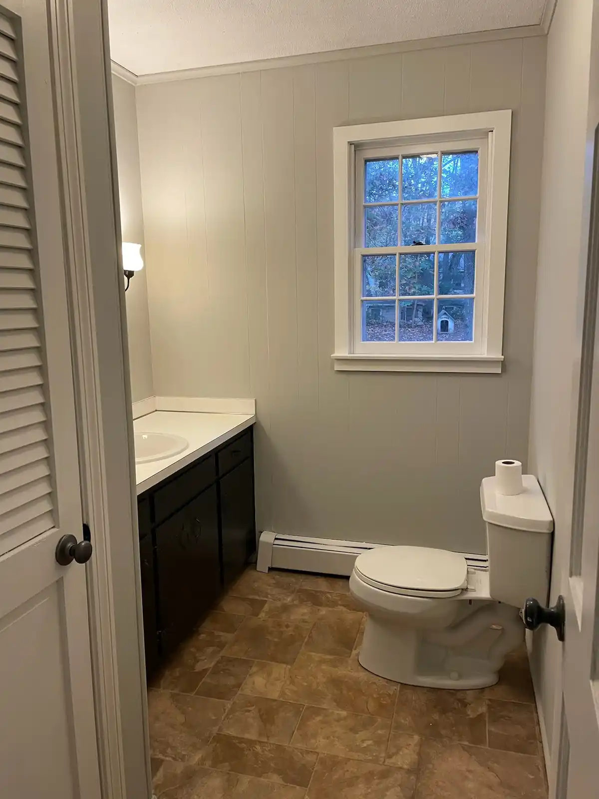 A clean, modern bathroom with a white toilet, dark vanity, and tiled floor, viewed from the doorway. Through the window, a…