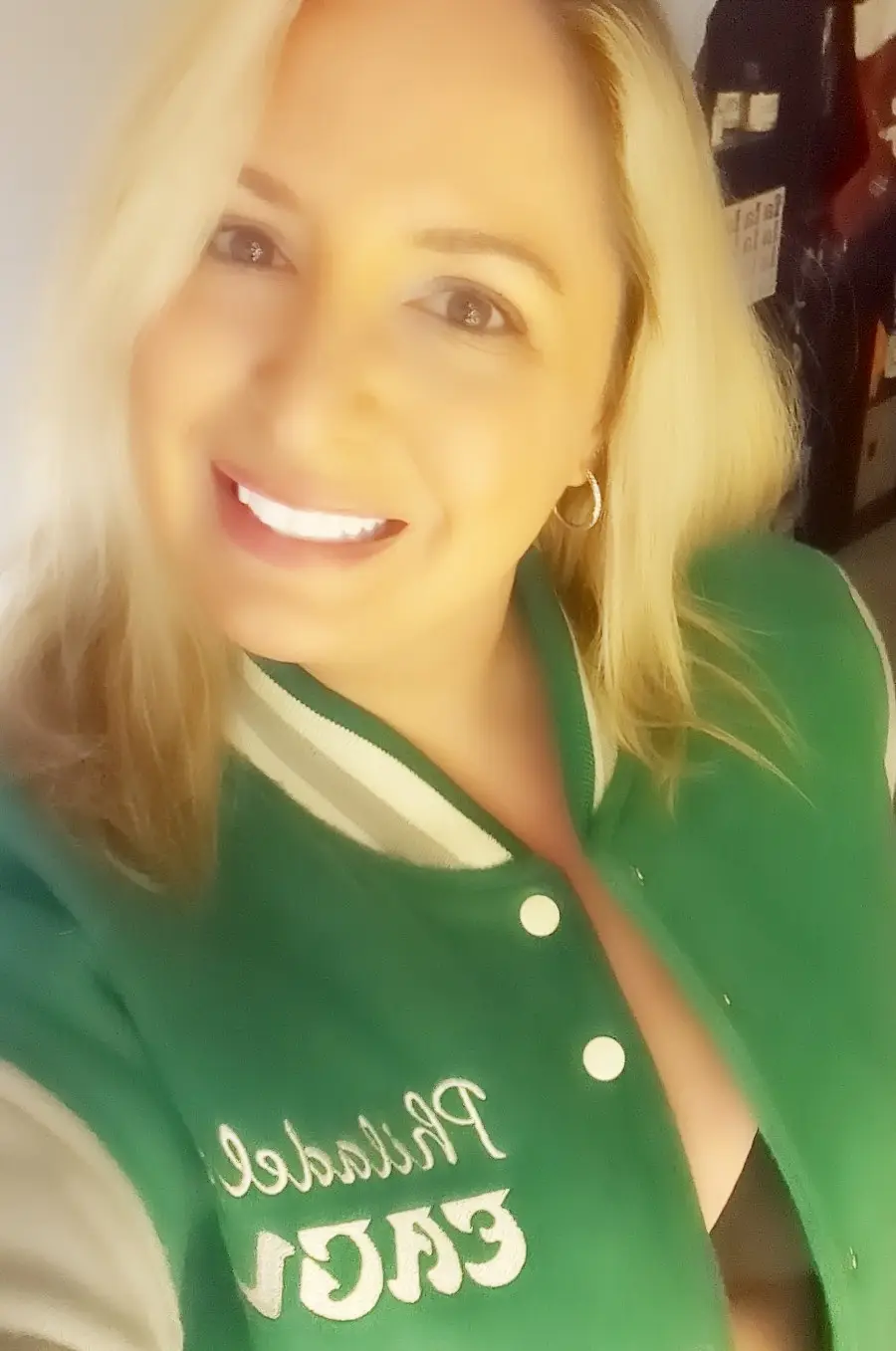 A smiling blonde woman in a green varsity jacket with white lettering that reads 'philadelphia eagles' on the chest. The…