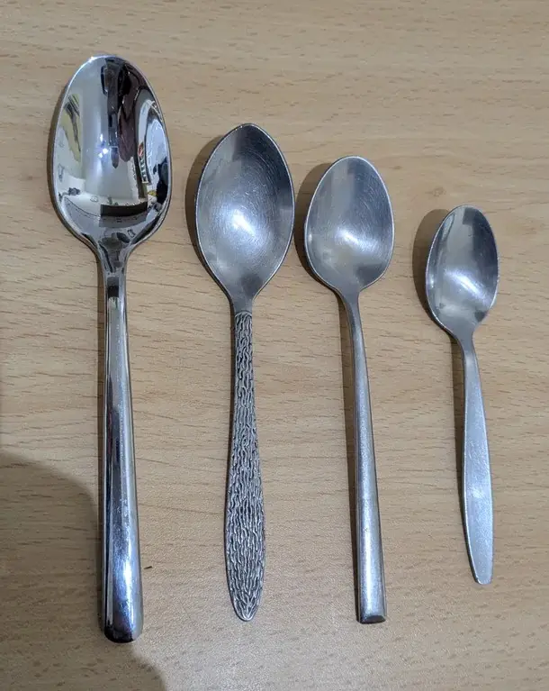 Four metal spoons of decreasing size are lined up on a light wood surface, with the largest on the left and smallest on the…