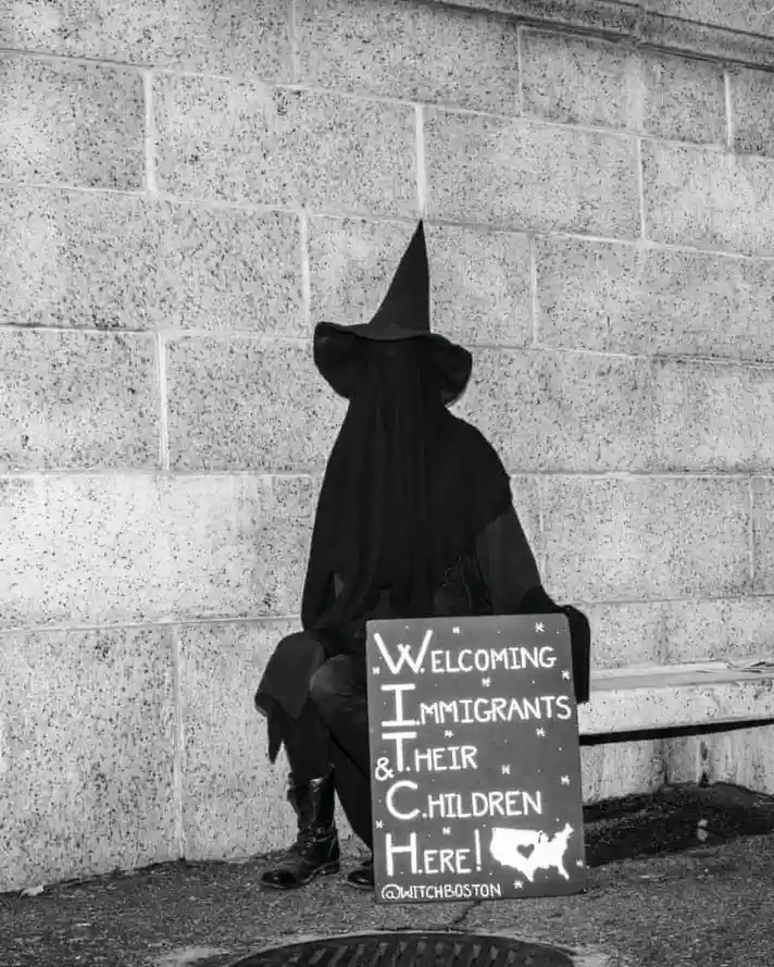 A person in a full black witch costume, including a pointed hat and veil, sits on a bench against a stone wall. They hold a…