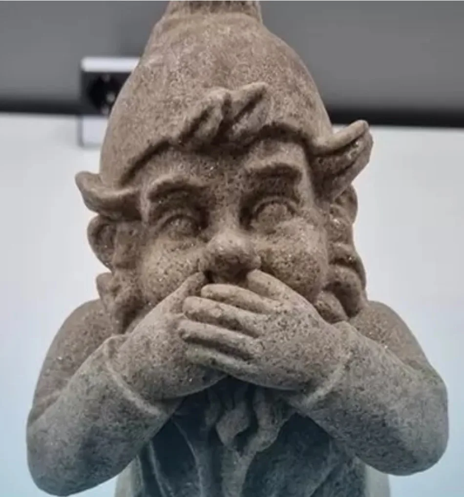 A close-up of a stone garden gnome statue with a surprised or shocked expression, covering its mouth with both hands. The…