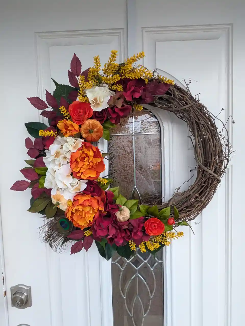 A colorful autumn-themed wreath made of artificial flowers, leaves, and small pumpkins hangs on a white front door with a…