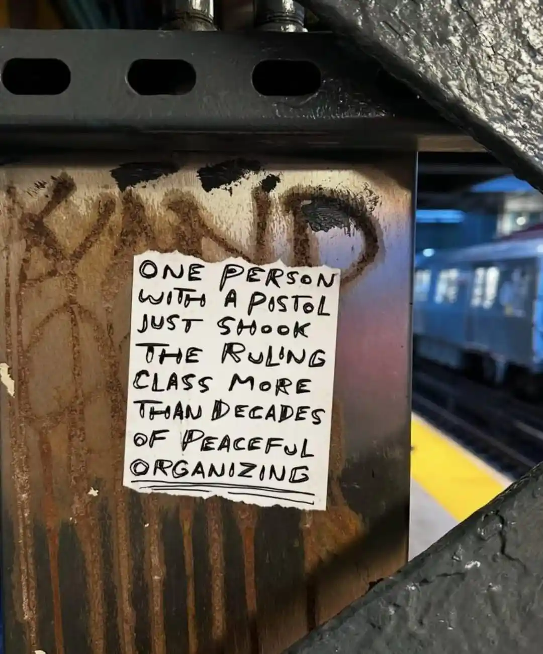 A handwritten note on a torn piece of paper is taped to a rusty metal pole in a subway station, with graffiti visible behind…