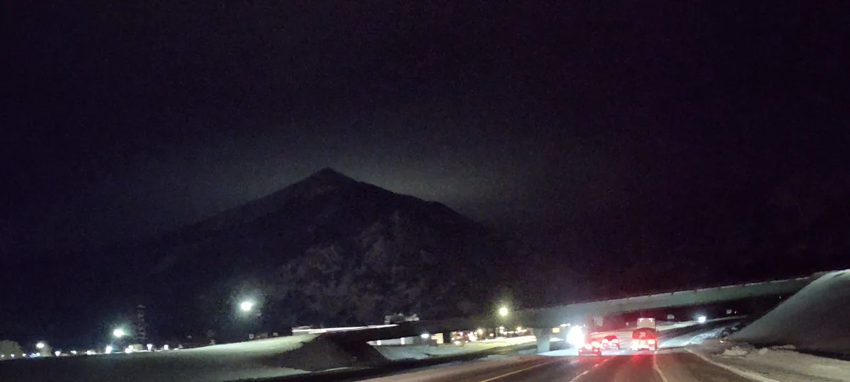 A nighttime photo taken from a car on a snowy highway, showing taillights of other vehicles ahead and a large, dark mountain…