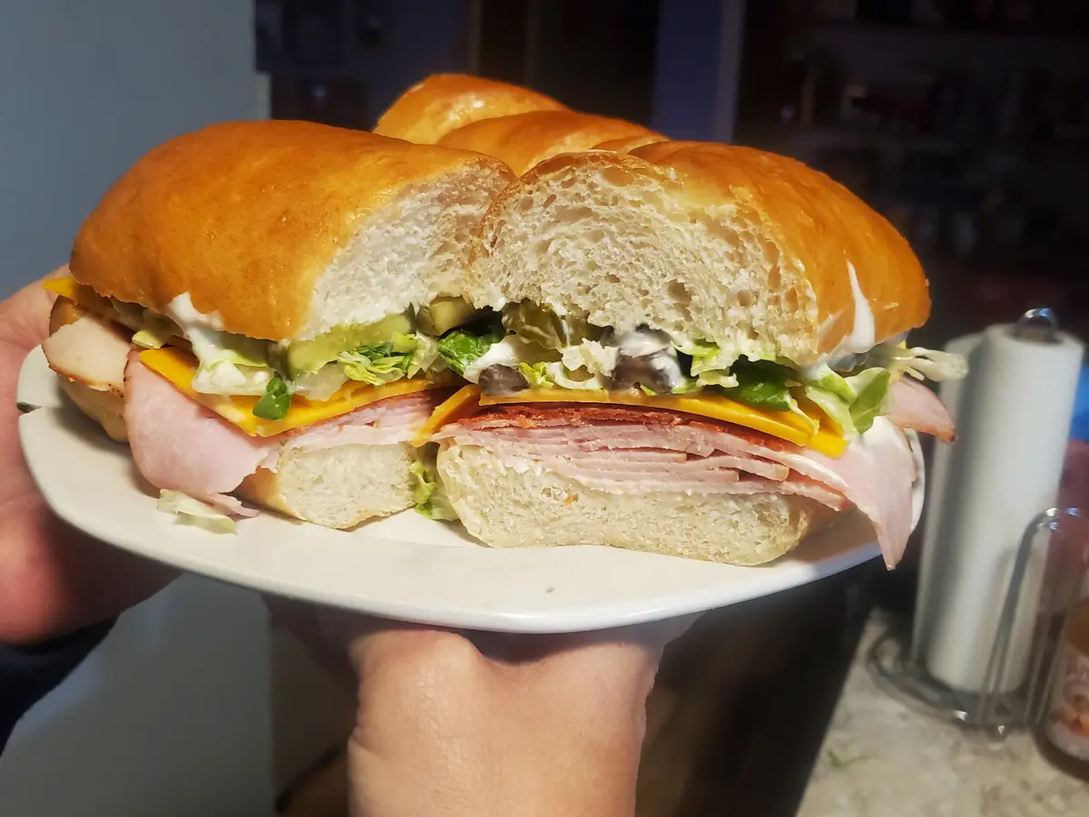 A person holds a white plate with a large, sliced sandwich in a soft roll, revealing layers of ham, cheese, lettuce…