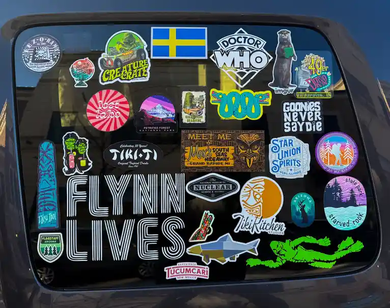 The image shows a car's rear window covered in numerous colorful stickers, including a Swedish flag, Doctor Who, Goonies…