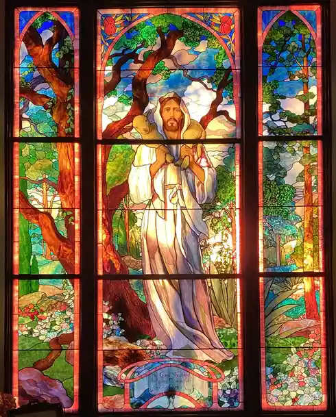 A stained glass window depicts Jesus Christ as the Good Shepherd holding a lamb, surrounded by trees and nature, with an…