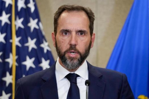 A man with a beard and dark suit stands at a podium with a microphone, looking shocked or alarmed. Behind him are two flags…