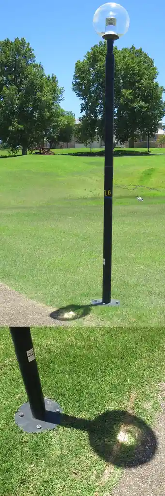 A black lamppost with a glass globe top stands in a grassy park under a clear blue sky. The pole has the number '16' painted…