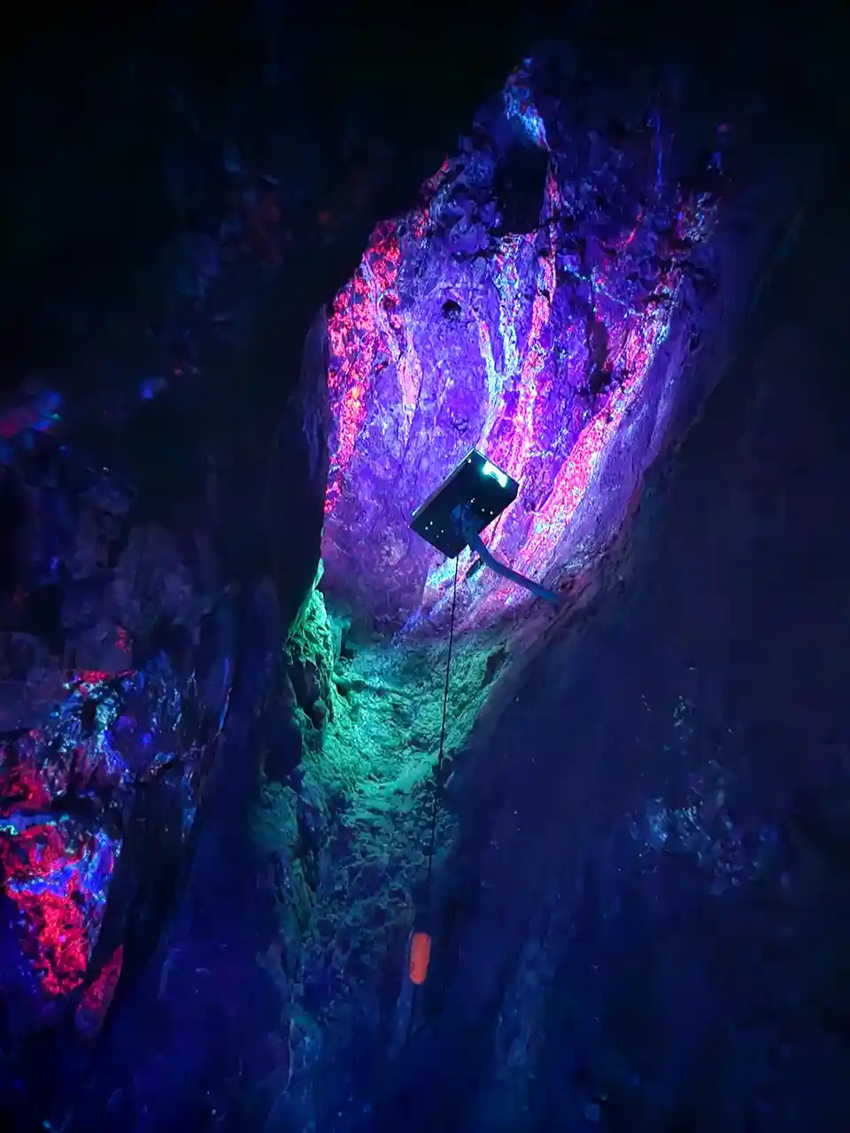 A dark cave interior is illuminated by vibrant purple and pink lights highlighting textured rock walls, with a small black…