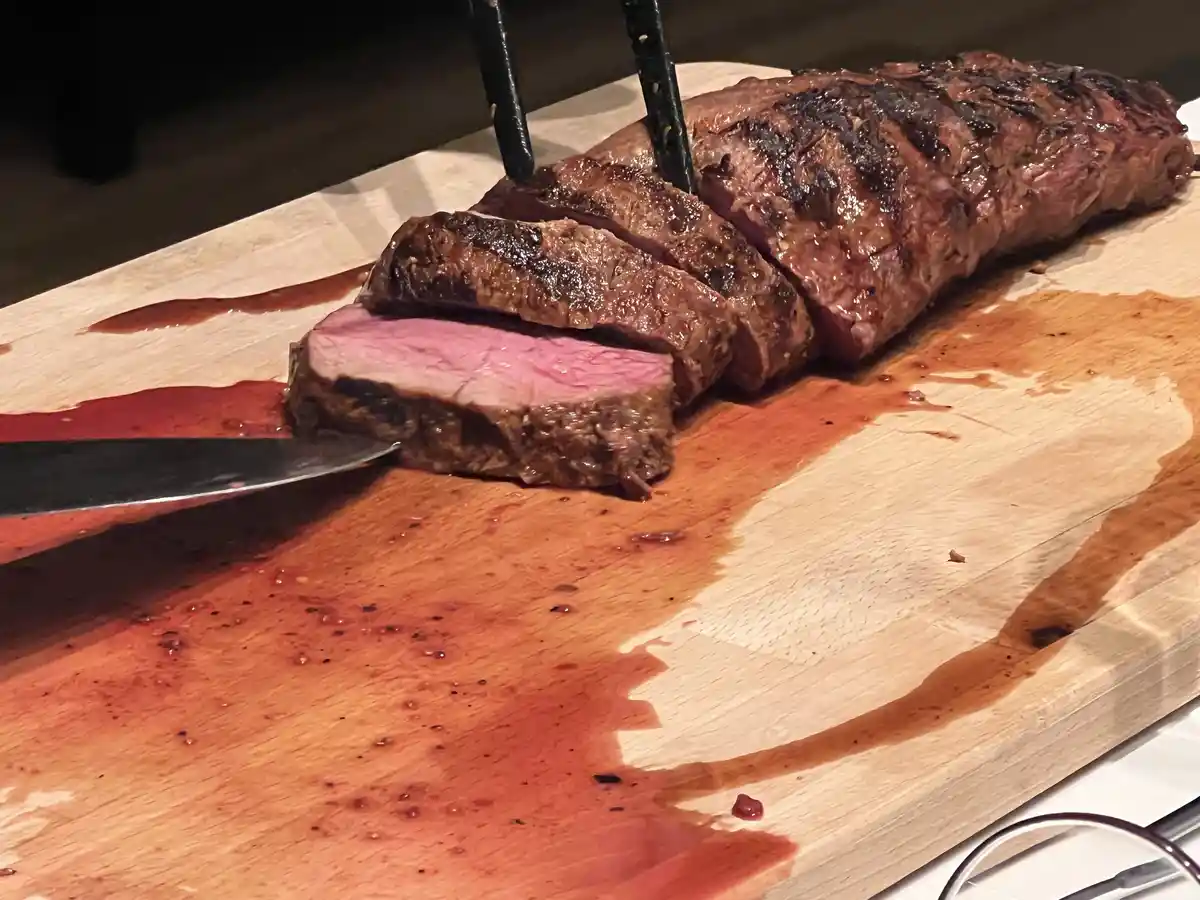 A close-up photo shows a large, grilled steak being sliced on a wooden cutting board, revealing a pink, rare interior. A…